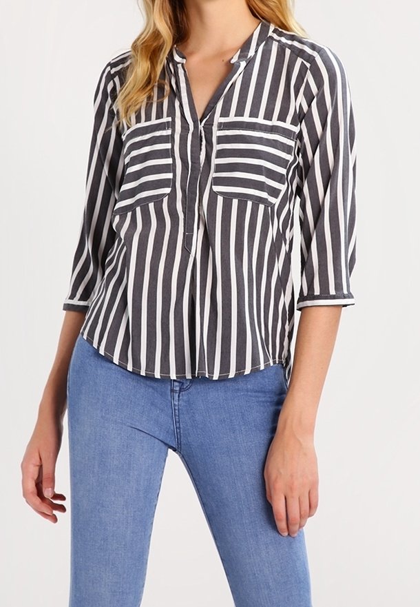 Femme portant un chemisier rayé vertical noir et blanc avec des manches 3/4 et deux poches poitrine, associé à un jean bleu.