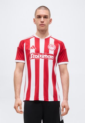 Muž nosi červeno-biely vertikálne pruhovaný futbalový dres Olympiacosu s logom sponzora Stoiximan, stojaci na bielom pozadí.