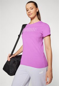 Kratkouruki vijoličen T-shirt s beleǰim logotipom "PUMA", v kombinaciji s svetlo sivo hlačami in črno torbo za šport, ki ima mrežni stranski del.