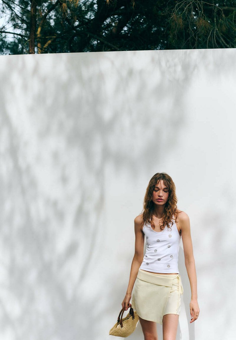 Femme aux cheveux bouclés portant un débardeur blanc et une jupe beige, tenant un sac tressé, marchant près d'un mur blanc avec des ombres d'arbres.