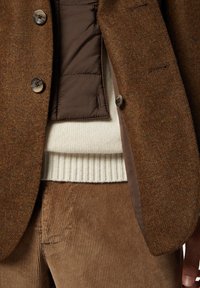 Giacca di tweed marrone con due bottoni, indossata sopra un maglione a coste crema e pantaloni in velluto a coste marroni. Un insieme testurizzato e casual.