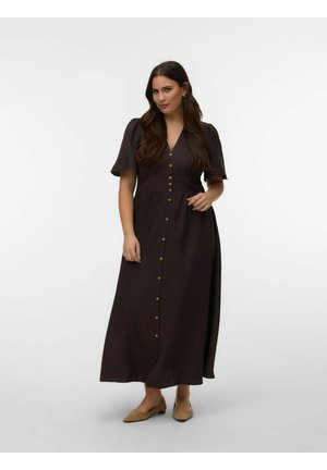 Moterys stovi dėvėdama tamsiai rudą mygtukais segamą midi suknelę su trumpomis pūstomis rankovėmis ir smėlio spalvos lygiais batais ant vienspalvio fono.