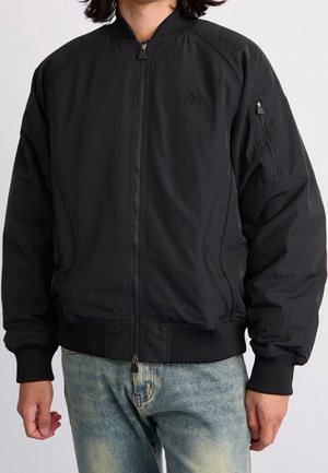 Personne portant une veste bomber Adidas noire avec une poche zippée sur la manche et un jean bleu clair.