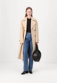 Beige trenchcoat med dobbelspent design og stor krage, kombinert med blå høytlivsjeans og svarte spisse støvler. Svart håndveske.