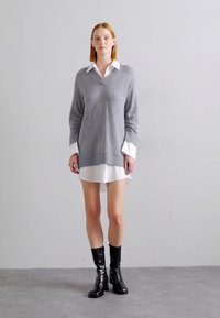 Pulover gri supradimensionat cu decolteu în V, purtat peste o bluză albă cu guler. Asortat cu cizme negre, lucioase, până la genunchi. Fundal uniform.