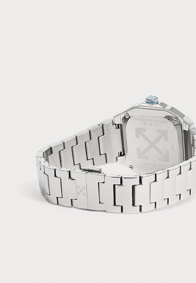 Montre-bracelet en métal argenté avec bracelet à maillons, montrant le dos gravé avec la résistance à l'eau et les marques de la marque.