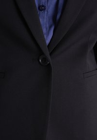 Svart blazer med enkel knappstängning, skräddarsydd passform och två sidofickor. Matchat med en blå skjorta med krage under.