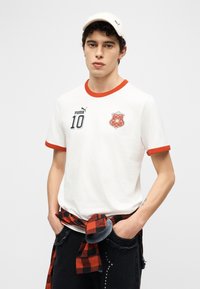 Νέος άντρας που φοράει λευκό μπλουζάκι Puma με  κόκκινη μπορντούρα, λευκό καπέλο και κόκκινο-μαύρο καρό πουκάμισο δεμένο γύρω από τη μέση του.