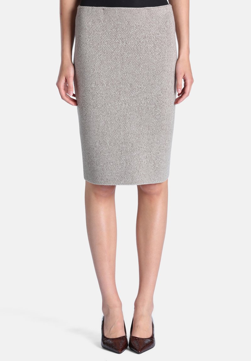 Femme portant une jupe crayon grise à chevrons jusqu'aux genoux et des talons hauts pointus foncés, debout avec les mains détendues le long du corps.