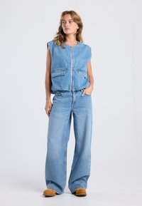 Light blue denim vest with a front zipper, two large pockets, paired with wide-leg light blue jeans and brown suede shoes.