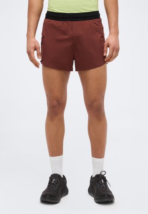 Mann trägt dunkelrote Sportshorts mit schwarzem Bund, weiße Sportsocken und schwarze Laufschuhe, steht vor einem schlichten Hintergrund.