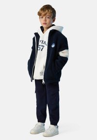 Niño pequeño vestido con una sudadera blanca, una chaqueta de forro polar azul marino con rayas blancas, pantalones cargo azul marino y zapatillas blancas, de pie con las manos en los bolsillos.