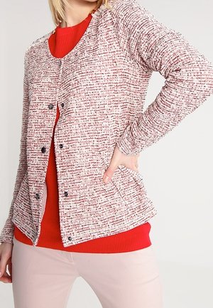 Kvinde iført en tekstureret lyserød og hvid knappet cardigan over en lys rød sweater, med lyserøde bukser, poserer med hånden på hoften.