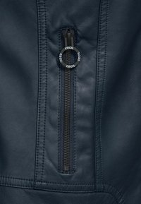 Szczegół marynarskiej skórzanej kurtki z gładką fakturą, zbliżenie na zamek błyskawiczny z okrągłym uchwytem oznakowanym "CECIL" oraz kontrastowym przeszyciem.