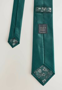 Teal satin slips med et blomstermønster på undersiden. Har en hexagonal spids og et label med teksten "NEXT Occasion" i guld.
