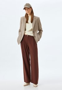 Brown wide-leg trousers paired with a cream sweater and a checked blazer. The model wears a matching checked cap and beige shoes.
