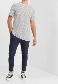 Krótki rękaw, prążkowana koszulka w białym i granatowym kolorze, z jednolitym blokiem kolorów na wysokości klatki piersiowej; w połączeniu z granatowymi, dopasowanymi spodniami i czarnymi trampkami.