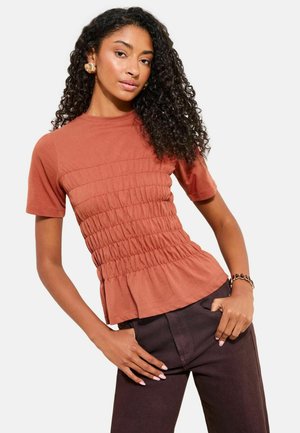 Žena s kudrnatými vlasy v rdzavě zbarveném nahuštěném tričku s krátkými rukávy a tmavých kalhotách pózující s jednou rukou v kapse a zlatými náušnicemi.