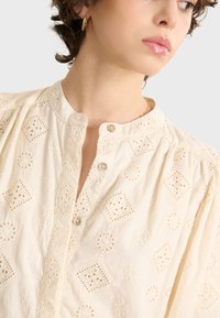 Crème-kleurige blouse met decoratieve borduurpatronen, ronde halslijn en knoopdetails. Lichtgewicht stof met een gestructureerde afwerking.