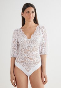 Witte kanten bodysuit met bloemenpatroon, doorschijnende stof, scalloped halslijn en opgeblazen mouwen. Effen witte onderrand als accent.