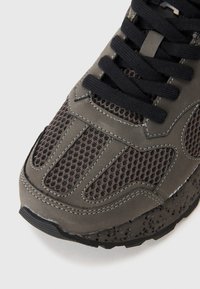 Gros plan de la partie avant d'une basket grise et noire avec des panneaux en mesh, des lacets noirs et une semelle texturée.