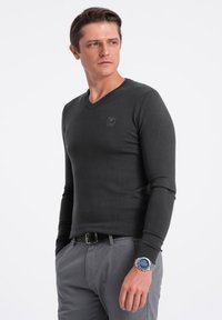 Maglione grigio scuro a maniche lunghe con scollo a V in tessuto morbido, con un piccolo logo sul petto. Abbinato a pantaloni grigi chiari e un orologio argento.