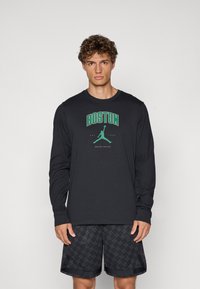 Sort langærmet t-shirt med "BOSTON" og et grønt grafik af en figur, der hopper. Båret med sorte mønstrede shorts.