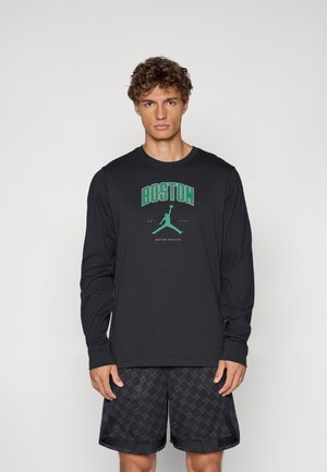 Sort langærmet t-shirt med "BOSTON" og et grønt grafik af en figur, der hopper. Båret med sorte mønstrede shorts.