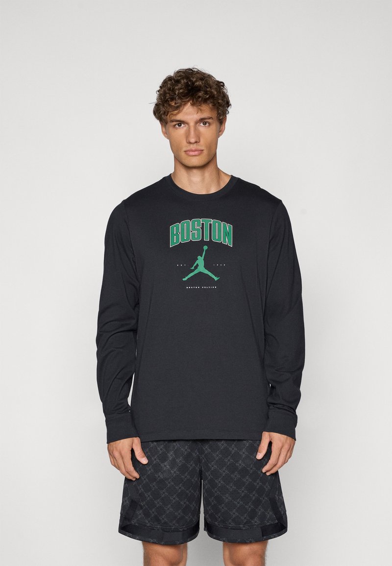 Sort langærmet t-shirt med "BOSTON" og et grønt grafik af en figur, der hopper. Båret med sorte mønstrede shorts.