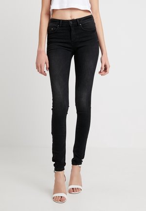 Czarne obcisłe jeansy noszone z białymi sandałami na wysokim obcasie z odkrytymi palcami oraz białym, krótkim topem, widoczne od talii do stóp.