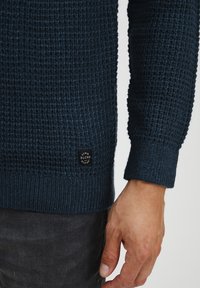 Námořnická modrá pletená mikina s texturovaným vzorem, žebrované manžety a černá značka na spodním levém rohu u lemování.