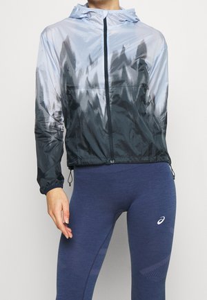 Frau trägt eine hellgraue und schwarze Kapuzenjacke mit Reißverschluss sowie blaue, eng anliegende Sportleggings mit einem weißen Logo am Oberschenkel.