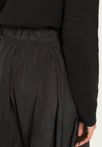 Fustă textilă neagră cu pliuri și talie elastică, asortată cu un pulover tricotat negru. Materialul are o textură netedă și lucioasă.