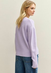 Vrouw met lang blond haar, gekleed in een lichtpaarse trui en blauwe jeans, staand in zijaanzicht tegen een eenvoudige lichte muur.