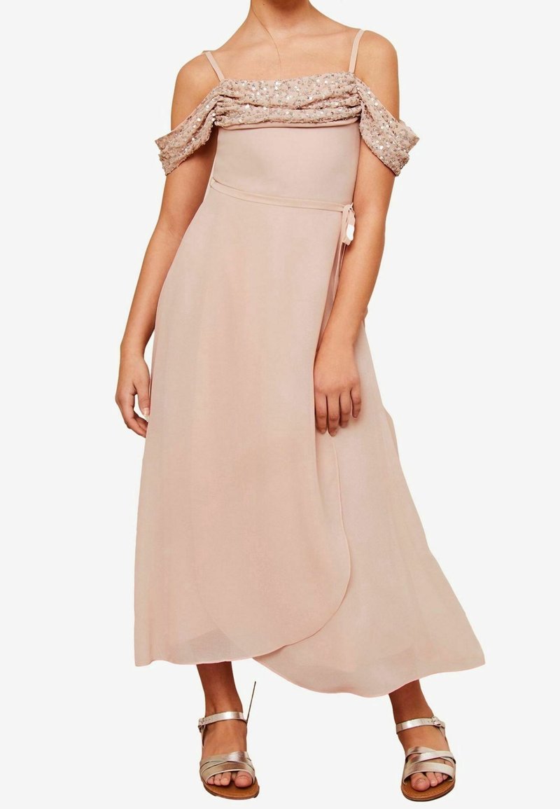 Femme portant une robe midi rose clair à épaules dénudées avec un tissu pailleté sur le haut de la poitrine, associée à des sandales métalliques à lanières.
