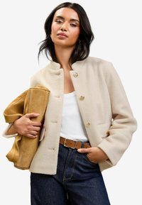 Beige uldblandet jakke med høj krave, guldknapper og frontlommer, parret med en tan ruskinds taske og højlife mørke denim jeans.