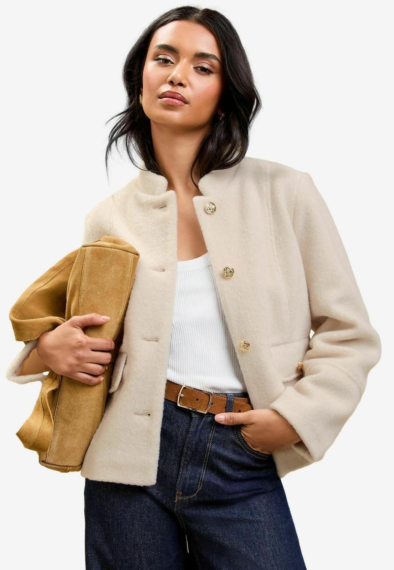 Beige uldblandet jakke med høj krave, guldknapper og frontlommer, parret med en tan ruskinds taske og højlife mørke denim jeans.