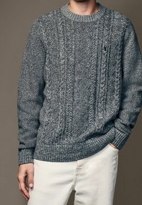 Suéter gris de punto grueso con patrones texturizados, cuello y puños acanalados, combinado con pantalones de color claro. Material suave y acogedor.