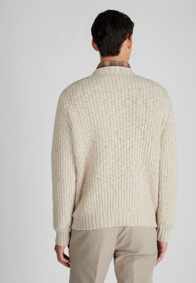 Cremfarbener Strickpullover mit strukturiertem Rippenmuster, mit hohem Kragen und lockerer Passform, getragen über einem karierten Hemd.