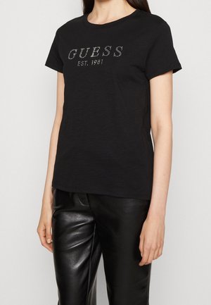 Woman wearing black short-sleeve shirt with "GUESS EST. 1981" in small studs and black leather pants against a plain light background.
