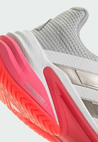 Zapato deportivo gris con parte superior de malla texturizada, acentos metálicos y una suela de goma rosa brillante con un patrón de banda de rodadura en zigzag.