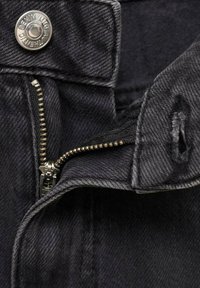 Tejido de denim negro con un cierre de plata visible y un botón a presión de metal. Presenta detalles de costura y una ligera textura. Ángulo en primer plano.