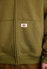 Casaco de fleece com fecho zip em verde-azeitona, com punhos e barra canelados. Apresenta um destacado destaque do logótipo "Dickies" na área do peito esquerdo.