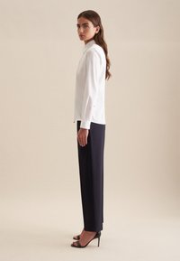 Camicia bianca a maniche lunghe con colletto, abbinata a pantaloni eleganti blu navy e sandali neri con tacco alto. Tessuto liscio senza motivi visibili.