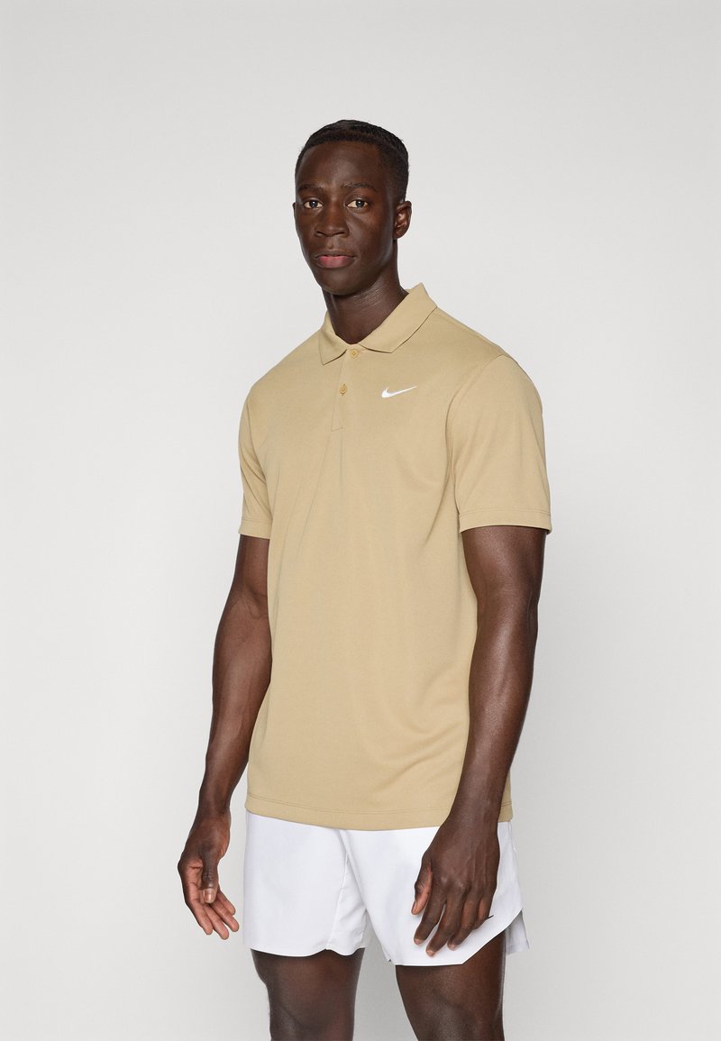 Beige Nike pólóing, galléros nyakkal, három gombos gombolással és fehér logóval. Fehér rövidnadrággal viselve, világos háttér előtt.