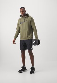 Sudadera verde oliva con logo negro de Nike, pantalones cortos negros y zapatillas negras. Sujetando una mancuerna, de pie contra un fondo claro y liso.
