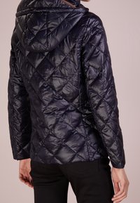 Marinblå kviltad pufferjacka med huva, som har ett diamantmönster och en slät, glansig yta. Dolda framfickor och en åtsittande silhuett.