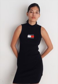 Abito senza maniche in maglia a costine nera, con un logo rettangolare "TOMMY JEANS" in rosso, bianco e blu nella zona del petto.