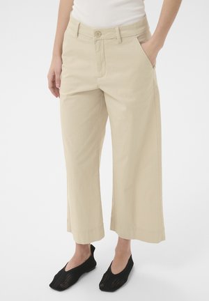 Pantaloni bej cu croială largă și tăietură scurtă purtați cu un top alb și pantofi negri împletiți slip-on, cu mâinile relaxate pe lângă corp.