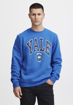 Kongelig blå hettegenser med "YALE" i fet hvit tekst, rund hals, ribbestrikkede mansjetter, og en logo med laurbærdesign i midten.